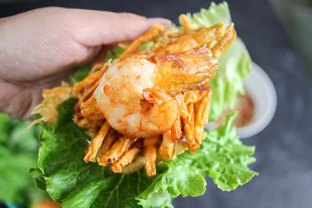 A hand holds a piece of green leaf lettuce with a Vietnamese Shrimp and Sweet Potato Fritters (Bánh Tôm Chiên Khoai Lang) inside.