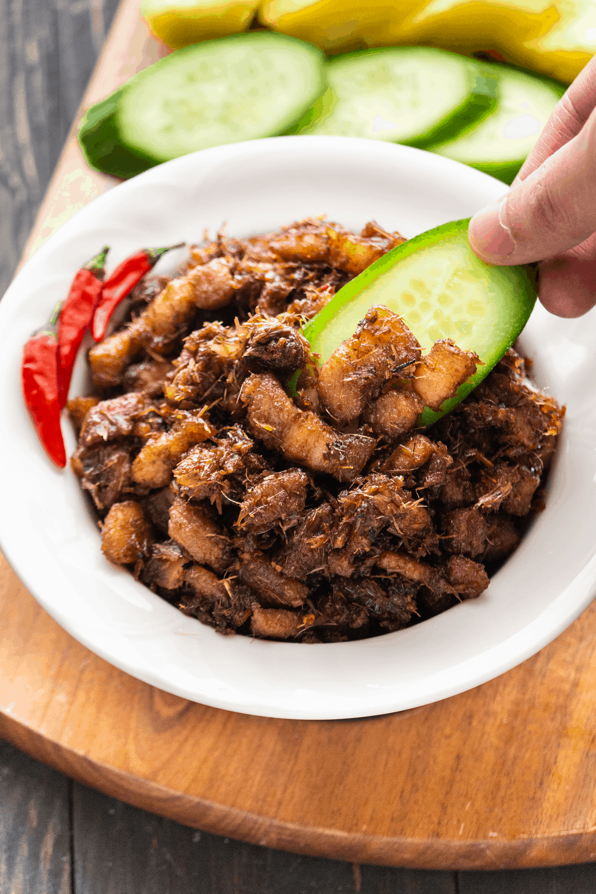 Thịt Kho Mắm Ruốc in a bowl with a hand dipping cucumber into it.