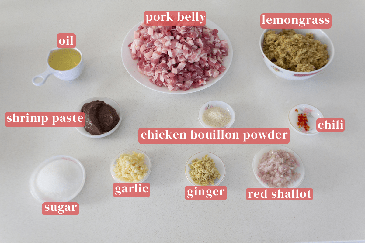 A plate of chopped pork belly along with bowls of finely chopped lemongrass and oil near dishes of chopped chili, red shallots, ginger and garlic with dishes of chicken bouillon powder, sugar and shrimp paste.