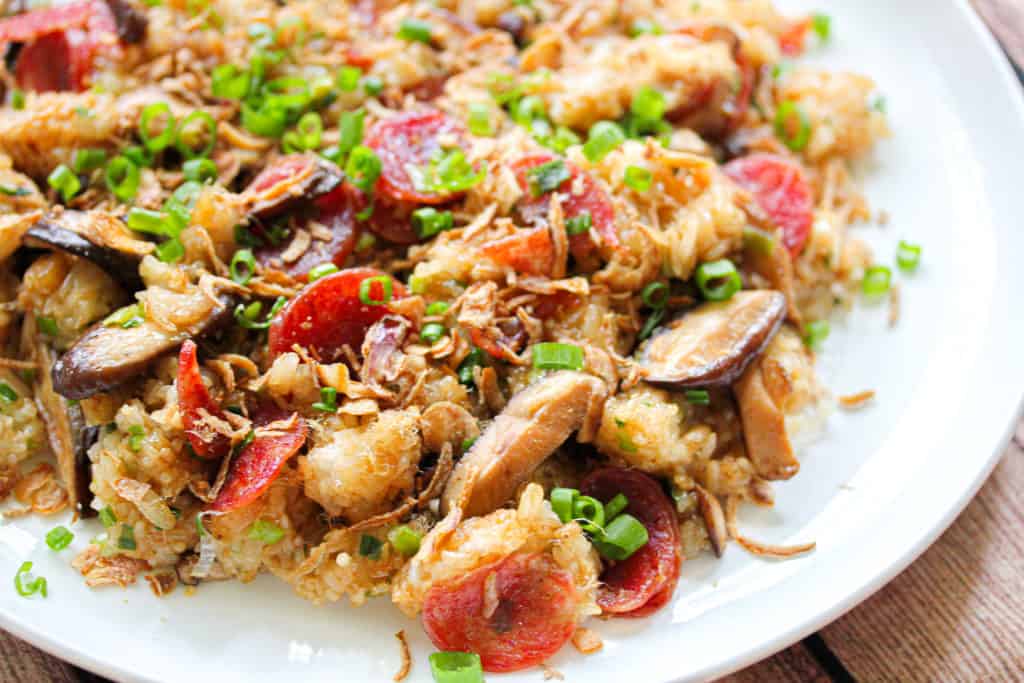 A close up shot partially showing a white plate. Brown sticky rice with chinese sausage, sliced mushroom and green onion are mixed through the rice