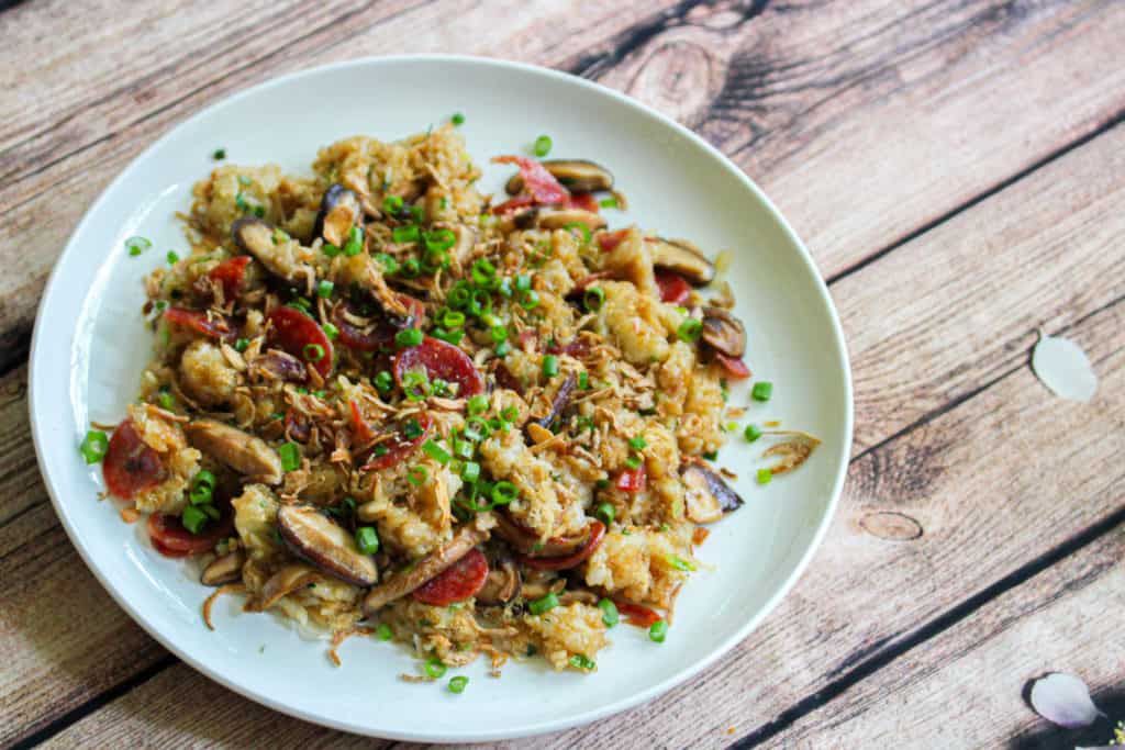 a white plate on on top of a fabric with a wooden pattern. On top of the plate is brown sticky rice with bits of chinese sausage, green onion and fried shallot mixed throughout.