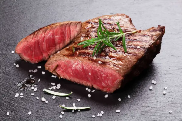 Một miếng steak bò nướng được cắt thành hai phần, trang trí với lá rosmarin và hạt muối trên bề mặt đá màu đen.