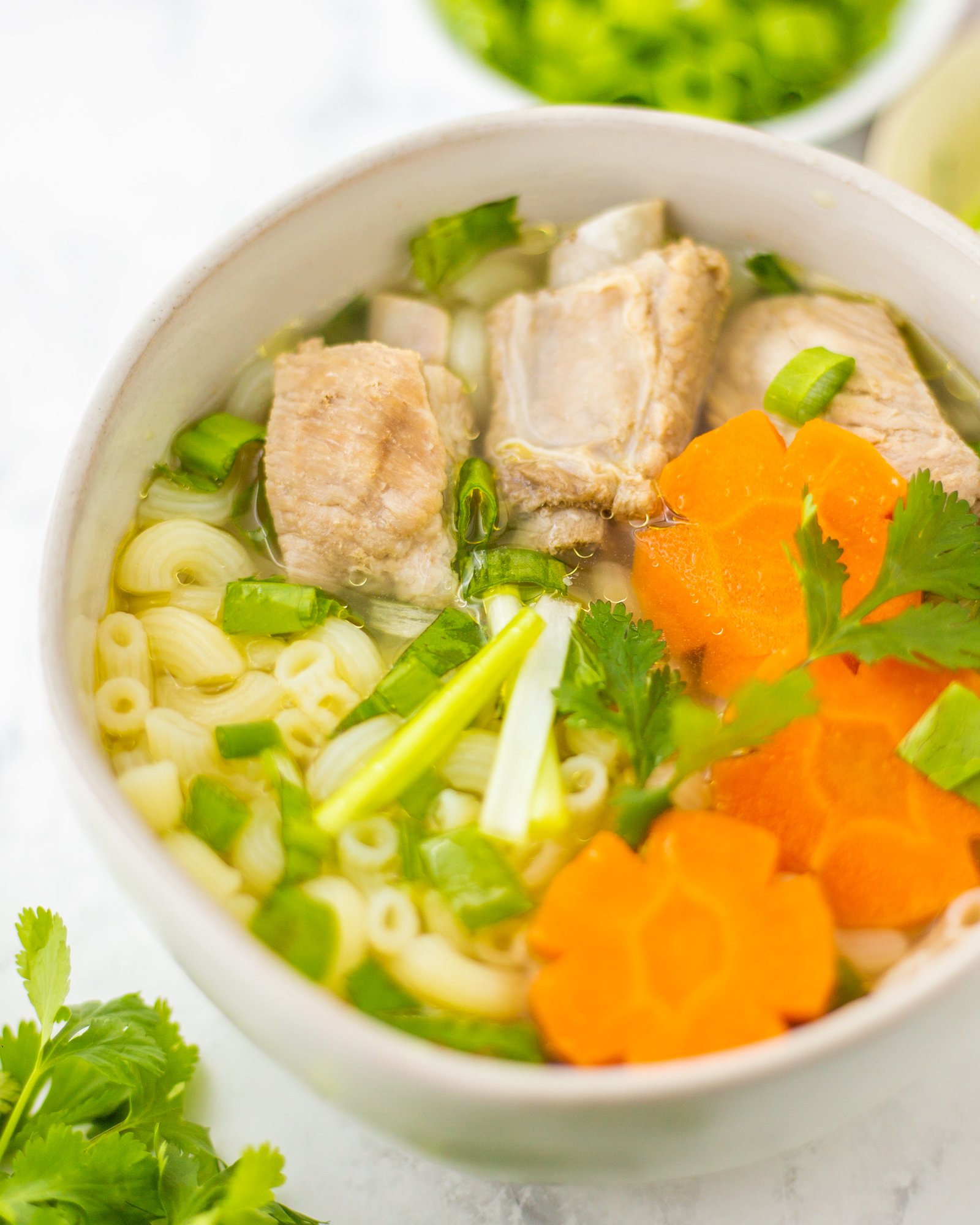 A white bowl containing macaroni pasta, ribs, slices of carrots, and some herbs in a clear broth.