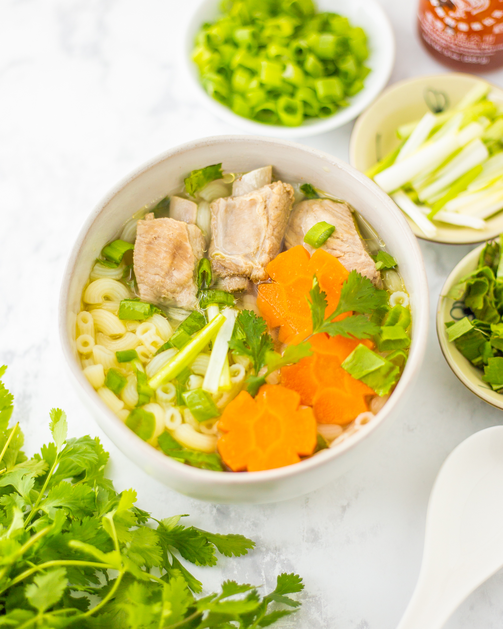 A white bowl containing macaroni pasta, ribs, slices of carrots, and some herbs in a clear broth.